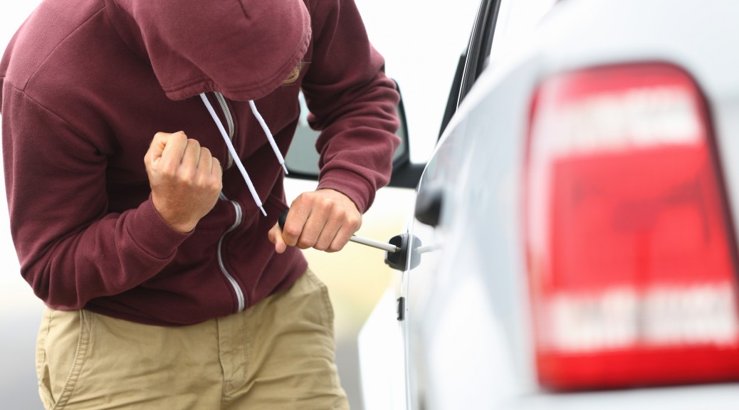 View down the side of a car to a man in a hooded top breaking into a car with a screwdriver in order to steal it